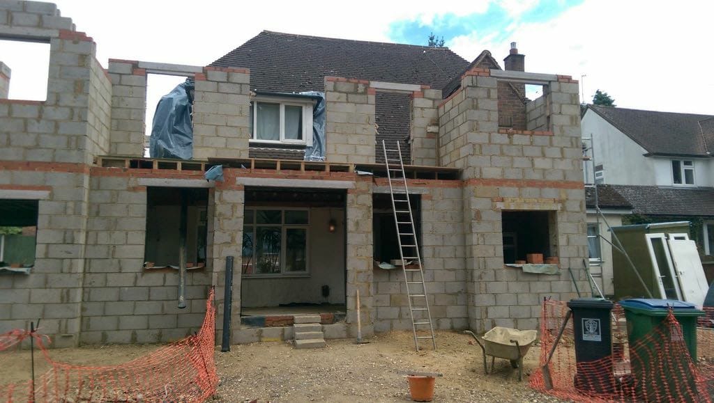 Budowa domu jednorodzinnego w trakcie realizacji: ściany z bloczków, widoczne otwory okienne i drzwiowe, drabina oparta o ścianę, teren budowy zabezpieczony siatką, w tle dom sąsiada.