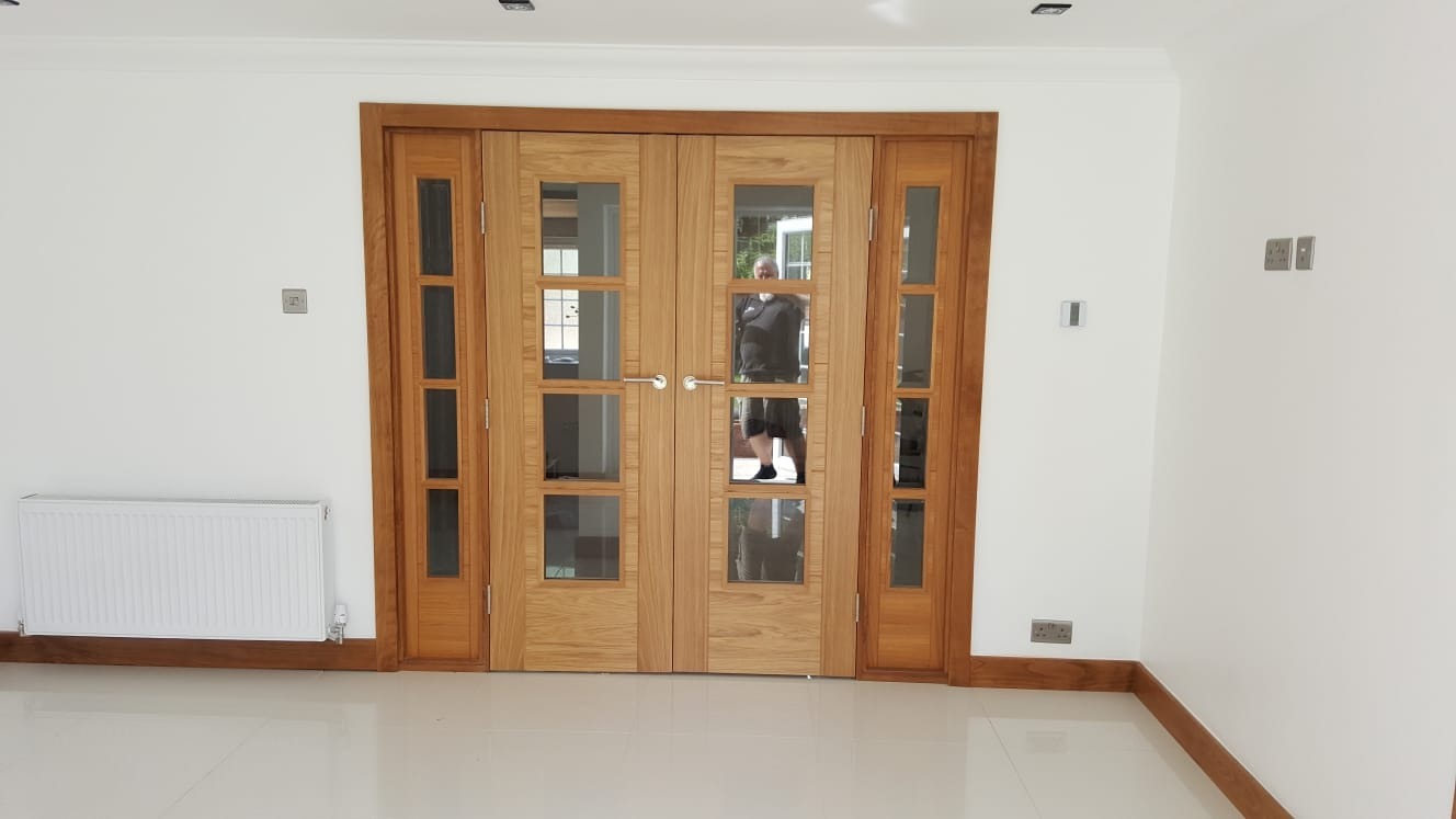 Nowoczesne, drewniane drzwi wewnętrzne z przeszkleniami w jasnym pomieszczeniu z białymi ścianami i błyszczącą, jasną podłogą. W szkle odbija się osoba robiąca zdjęcie.