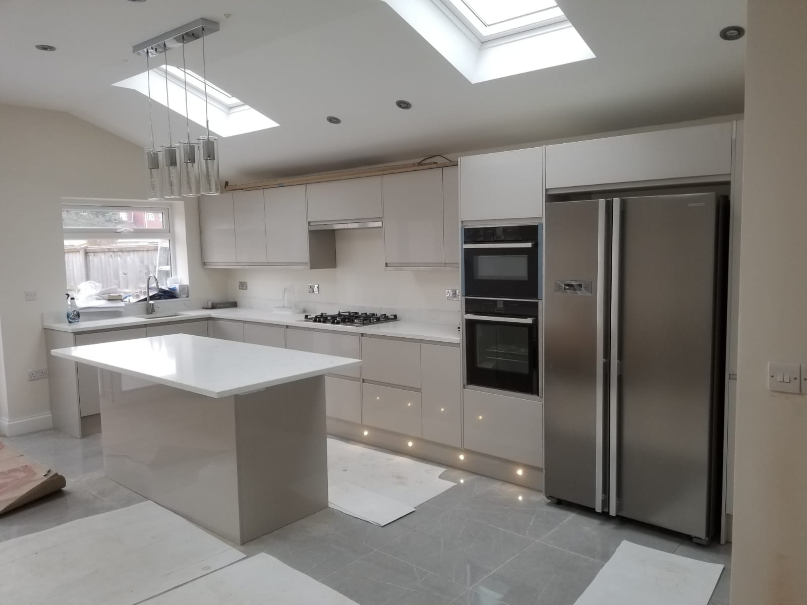 Nowoczesna kuchnia w odcieniach szarości z wyspą, oświetleniem podszafkowym i dużym oknem dachowym, na podłodze szare płytki, częściowo zabezpieczone kartonem.