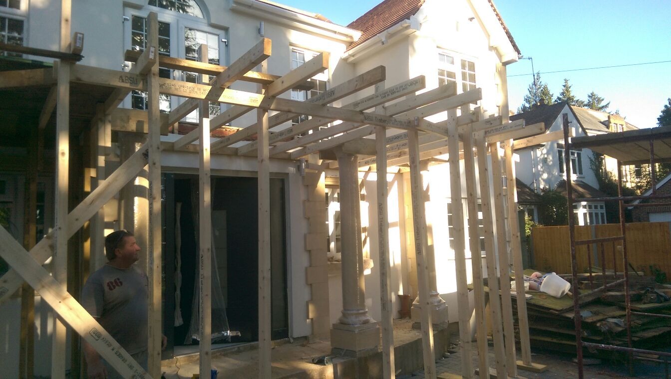 Budowa drewnianej konstrukcji zadaszenia tarasu przy domu jednorodzinnym, widoczny pracownik i elementy dekoracyjne elewacji.