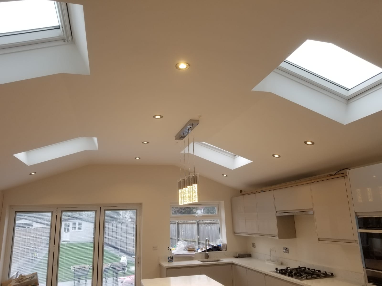 Jasna kuchnia z białymi szafkami, marmurowym blatem, oknami dachowymi i nowoczesnym oświetleniem wiszącym.