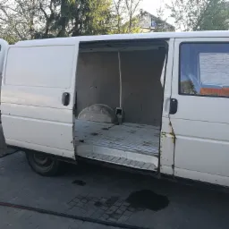 Biały, lekko zardzewiały bus dostawczy z otwartymi drzwiami bocznymi i tylnymi, ukazujący pustą przestrzeń ładunkową z metalową podłogą.