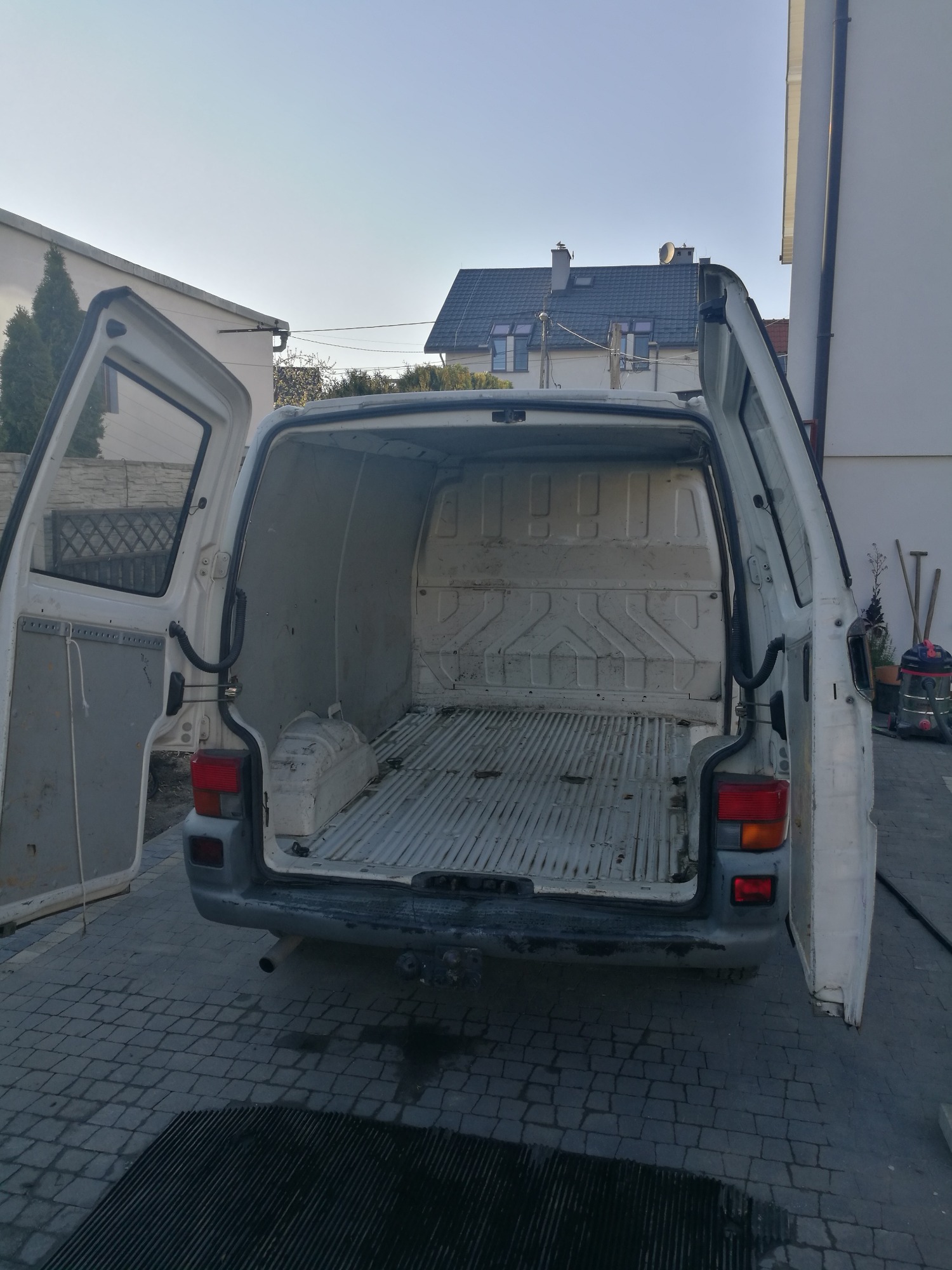 Wnętrze puste, białej furgonetki z otwartymi drzwiami, zaparkowanej na brukowanym podwórku przed budynkiem.