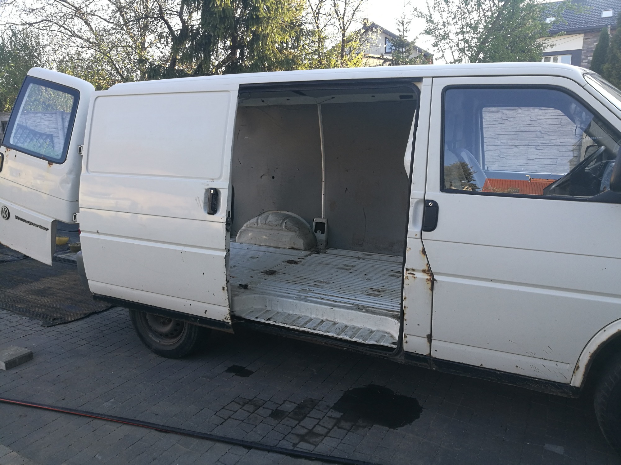 Biały, lekko zardzewiały bus dostawczy z otwartymi drzwiami bocznymi i tylnymi, ukazujący pustą przestrzeń ładunkową z metalową podłogą.