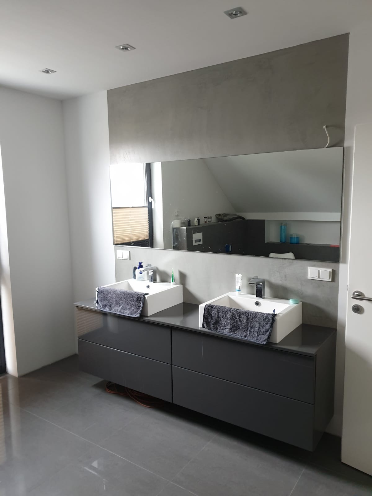 Nowoczesna łazienka z dwiema białymi umywalkami na szarej szafce, duże lustro na ścianie z szarą fakturą, szare płytki podłogowe.