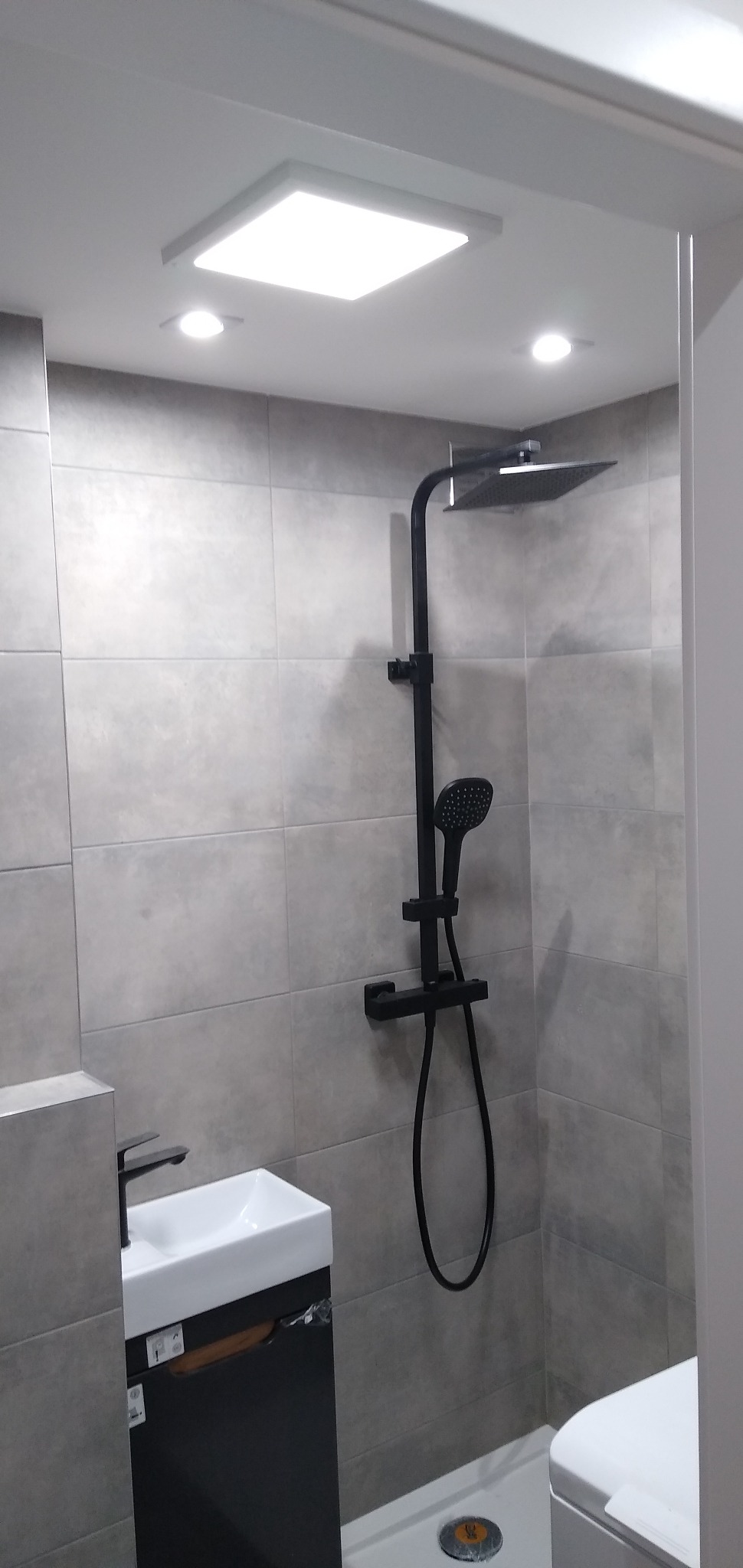 Wykończona łazienka z szarą, kwadratową glazurą, czarną armaturą prysznicową z deszczownicą i umywalką z szafką, oświetleniem sufitowym.