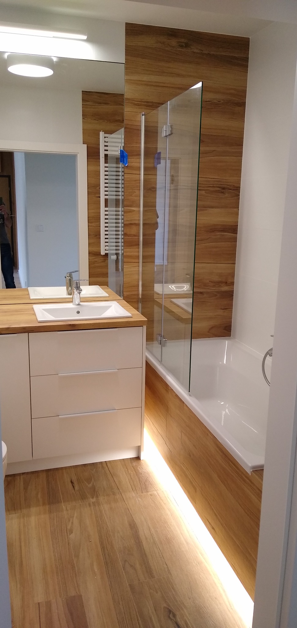 Nowoczesna łazienka z drewnopodobnymi płytkami, białymi meblami łazienkowymi z umywalką nablatową, parawanem nawannowym i podświetleniem LED pod wanną.