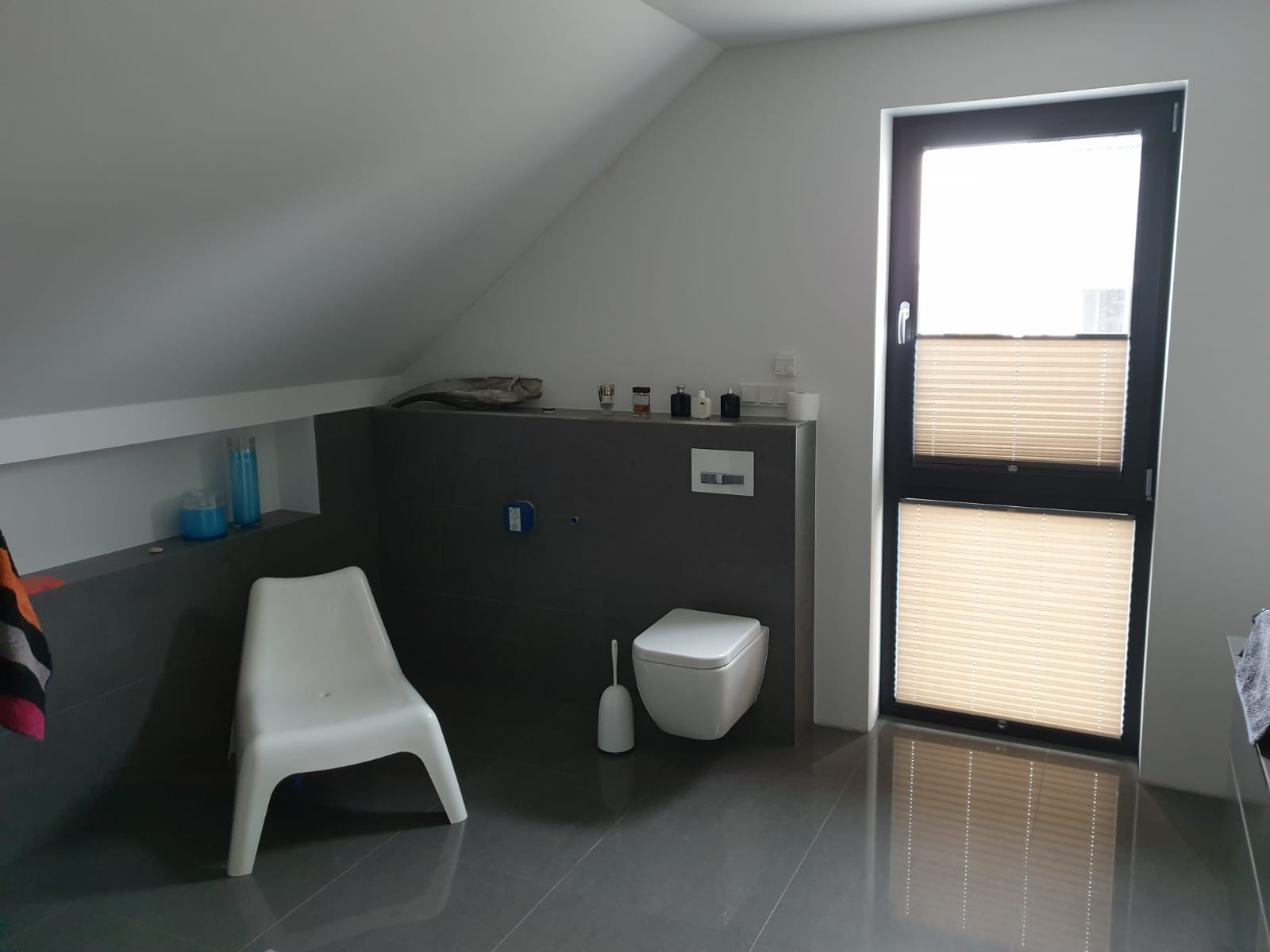 Nowoczesna łazienka na poddaszu z szarą glazurą, wiszącą toaletą, plastikowym krzesłem i oknem z plisowanymi roletami.
