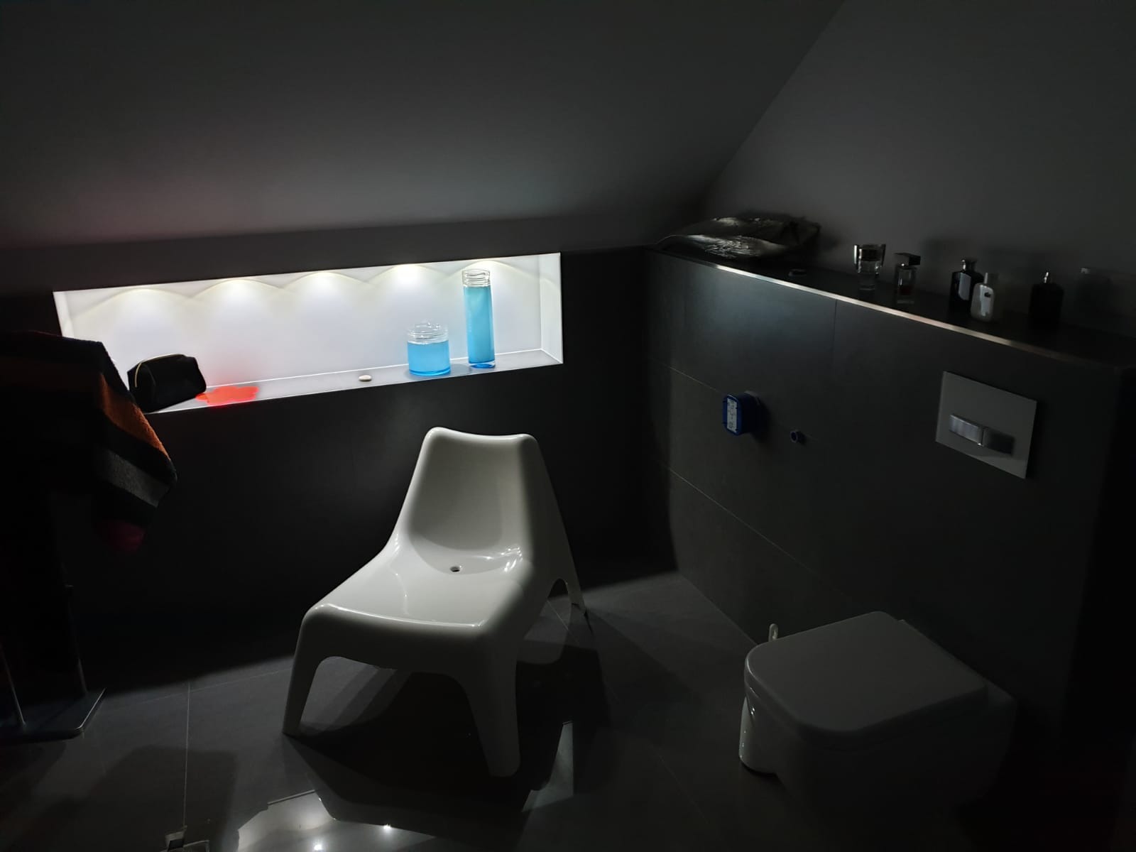 Ciemna łazienka na poddaszu z szarymi płytkami, białą miską WC, białą plastikową krzesłem i podświetlaną wnęką z dekoracjami w postaci szklanych pojemników z niebieską zawartością.