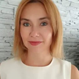 Portret kobiety z blond włosami i niebieskimi oczami, ubrana w białą bluzkę, na tle ściany z szarej cegły.