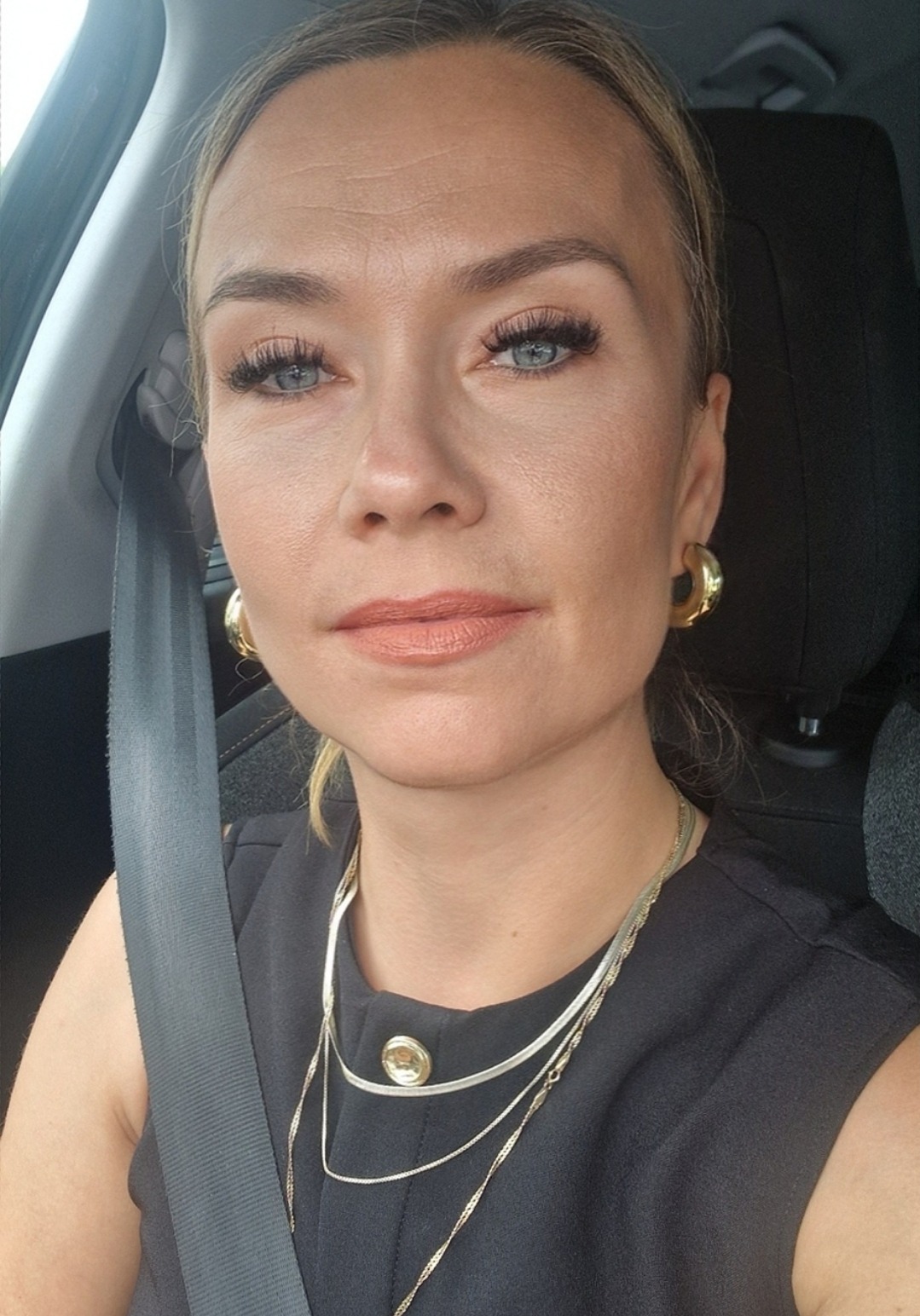 Portret kobiety z blond włosami, w czarnej bluzce i złotych kolczykach, siedzącej w samochodzie z zapiętym pasem bezpieczeństwa. Delikatny makijaż i srebrne łańcuszki.