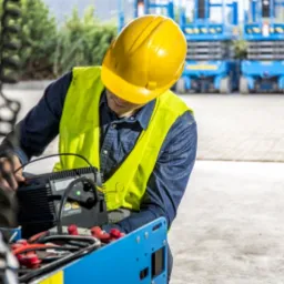 Pracownik w kasku i kamizelce odblaskowej w Zgierzu sprawdza prostownik do akumulatorów wózka widłowego, z tyłu widoczne inne wózki.