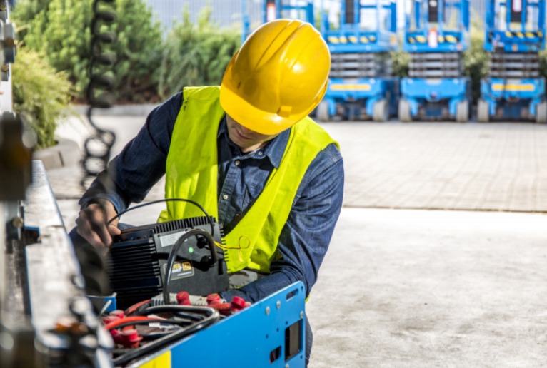 Pracownik w kasku i kamizelce odblaskowej w Zgierzu sprawdza prostownik do akumulatorów wózka widłowego, z tyłu widoczne inne wózki.