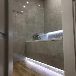 Nowoczesna łazienka z szarymi płytkami o geometrycznym wzorze, podświetleniem LED pod wanną i na ścianie, oraz drewnianą podłogą, widok zza otwartych drzwi.