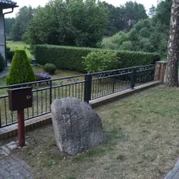 Czarne metalowe ogrodzenie z ceglanymi słupkami na tle zielonego żywopłotu i drzew, z dużą skałą z numerem domu '10' na pierwszym planie.
