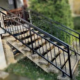 Zewnętrzne schody wejściowe z szarego betonu z czarną, metalową poręczą z dekoracyjnymi elementami, prowadzące do drzwi wejściowych, w tle zieleń.