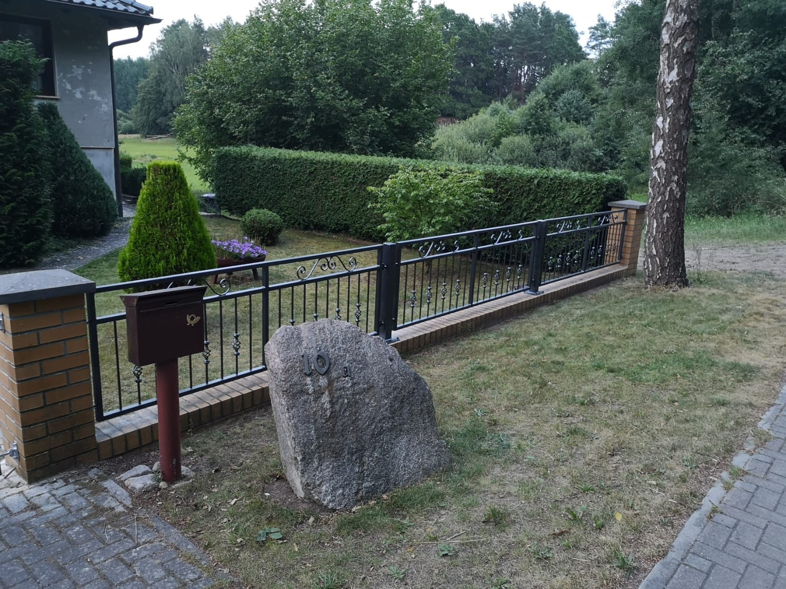 Czarne metalowe ogrodzenie z ceglanymi słupkami na tle zielonego żywopłotu i drzew, z dużą skałą z numerem domu '10' na pierwszym planie.