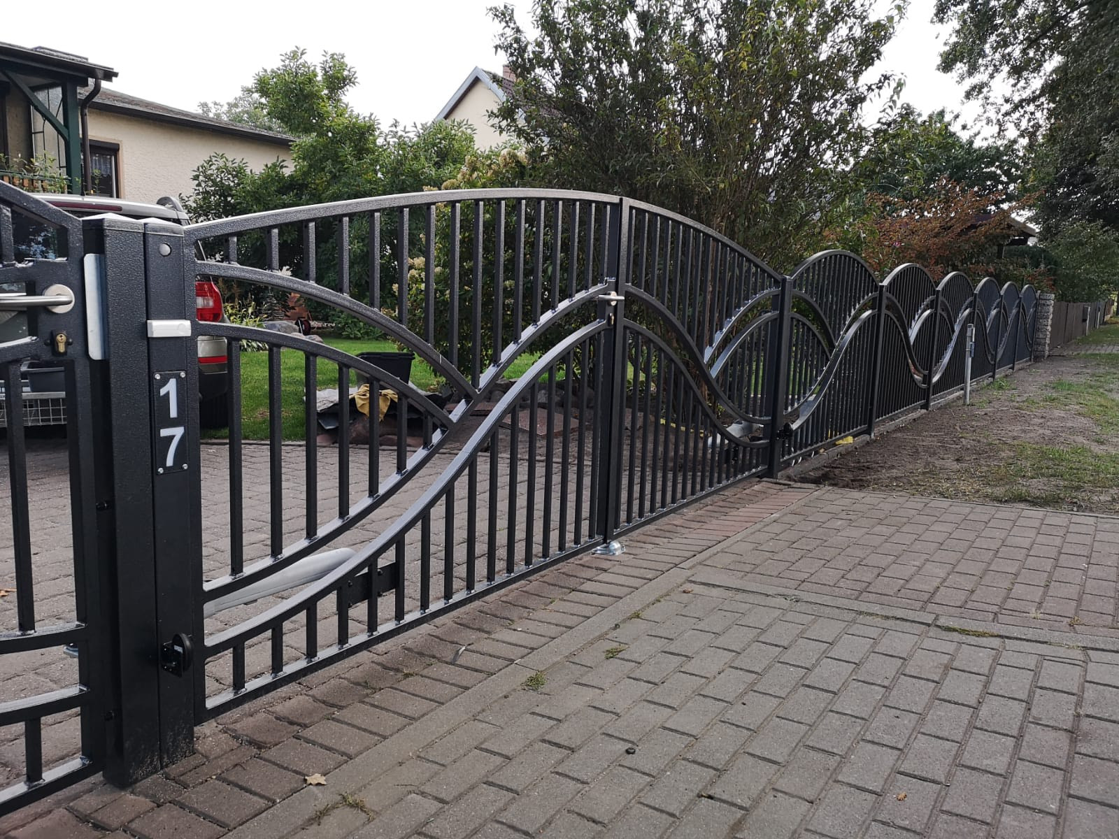 Czarna, metalowa brama wjazdowa z automatyką, numer posesji '17' widoczny na słupku, na tle kostki brukowej i ogrodzenia panelowego.