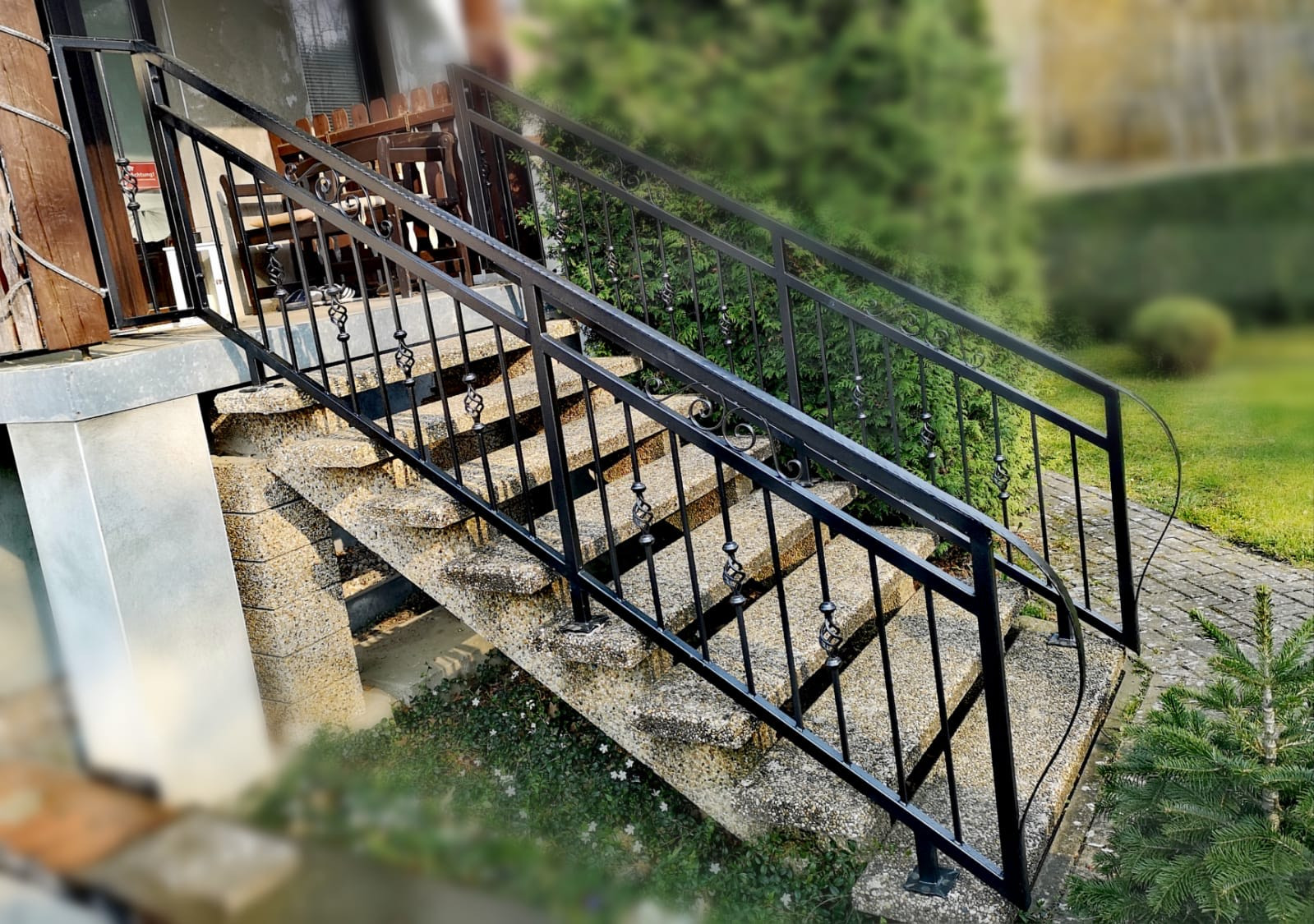 Zewnętrzne schody wejściowe z szarego betonu z czarną, metalową poręczą z dekoracyjnymi elementami, prowadzące do drzwi wejściowych, w tle zieleń.