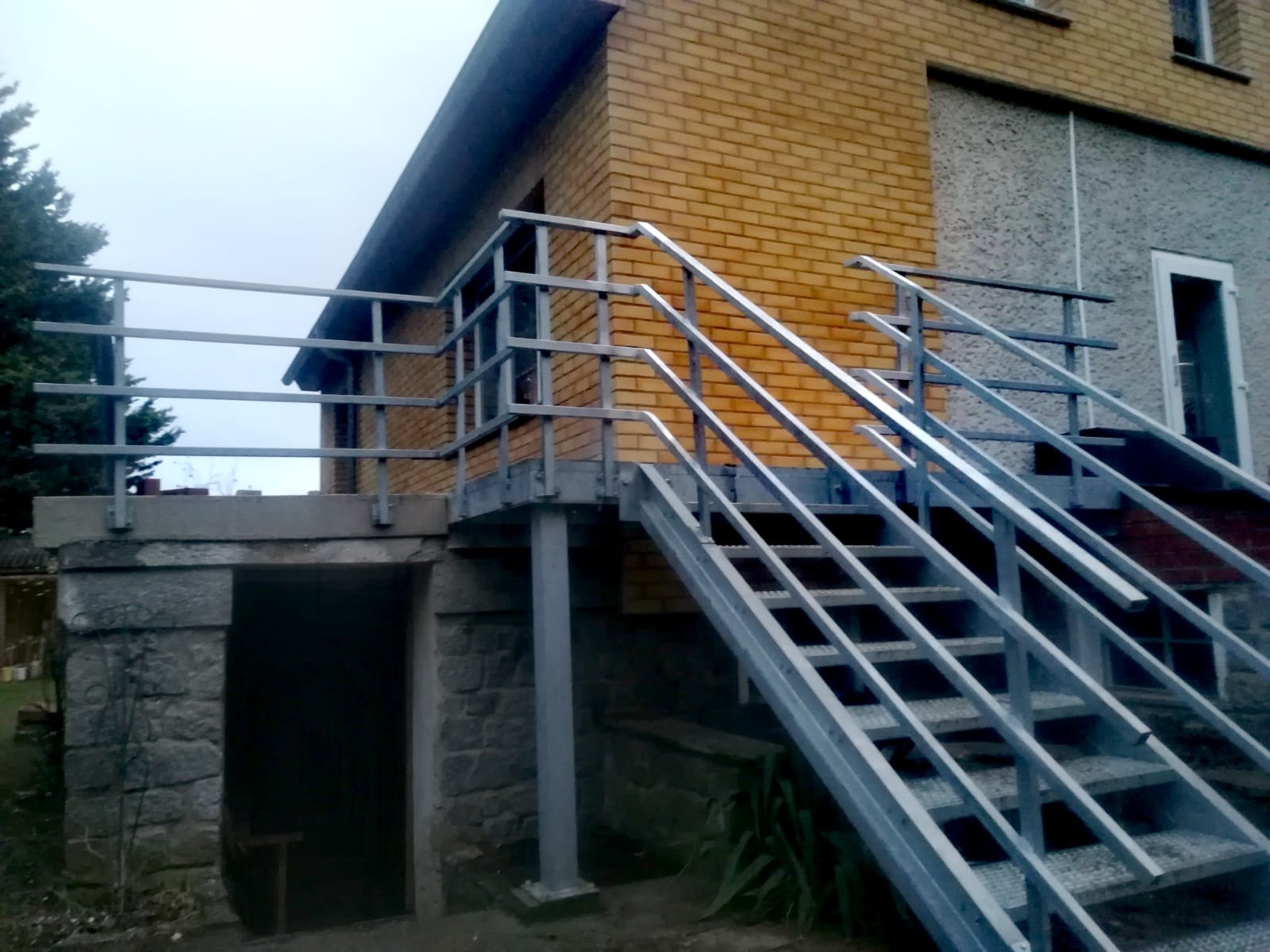 Zewnętrzne, metalowe schody z poręczami prowadzące do budynku z cegły i szarego tynku, widok z dołu, pochmurny dzień.
