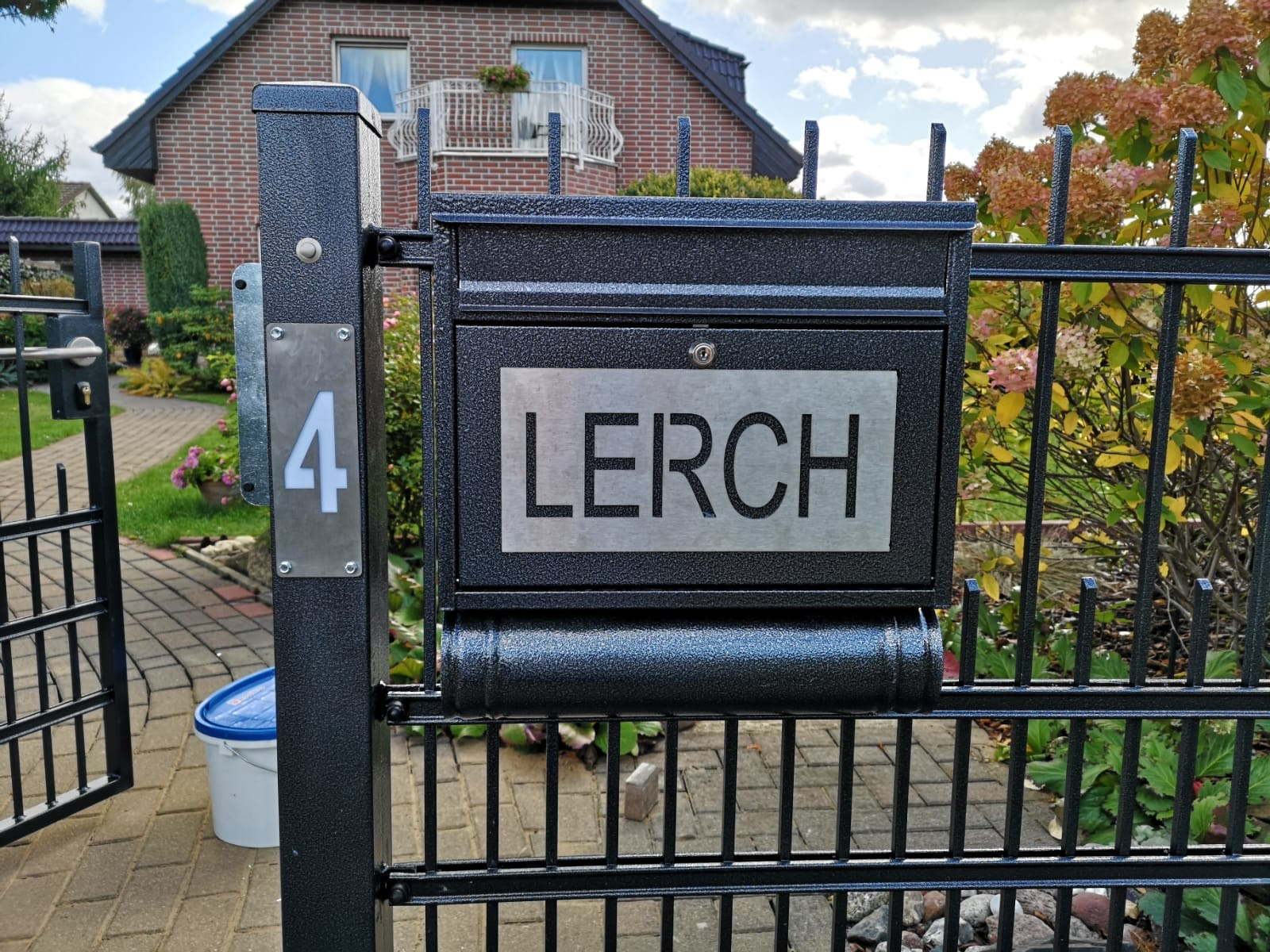Czarna metalowa furtka z numerem domu '4' i skrzynką pocztową z nazwiskiem 'LERCH', na tle domu z czerwonej cegły i roślinności.