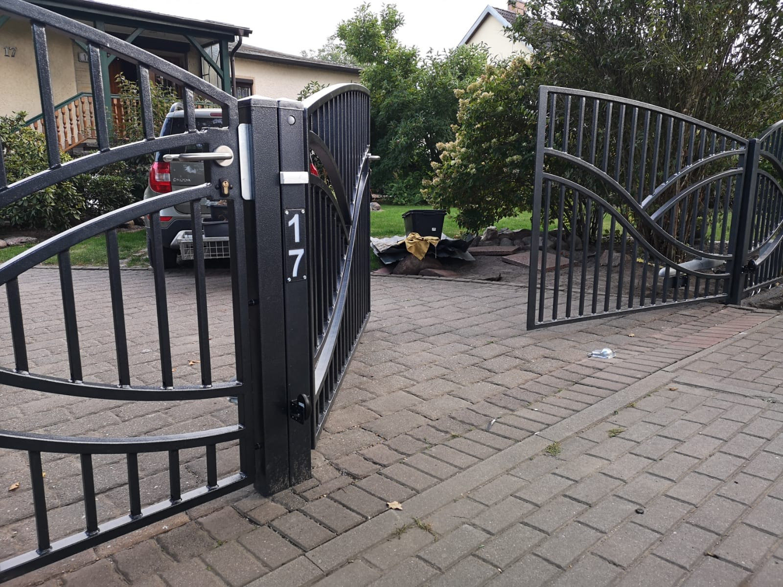 Otwarta, dwuskrzydłowa, metalowa brama wjazdowa w kolorze grafitowym z numerem posesji '17' na słupku, widoczna z poziomu brukowanej kostki, w tle częściowo widoczny samochód i dom.