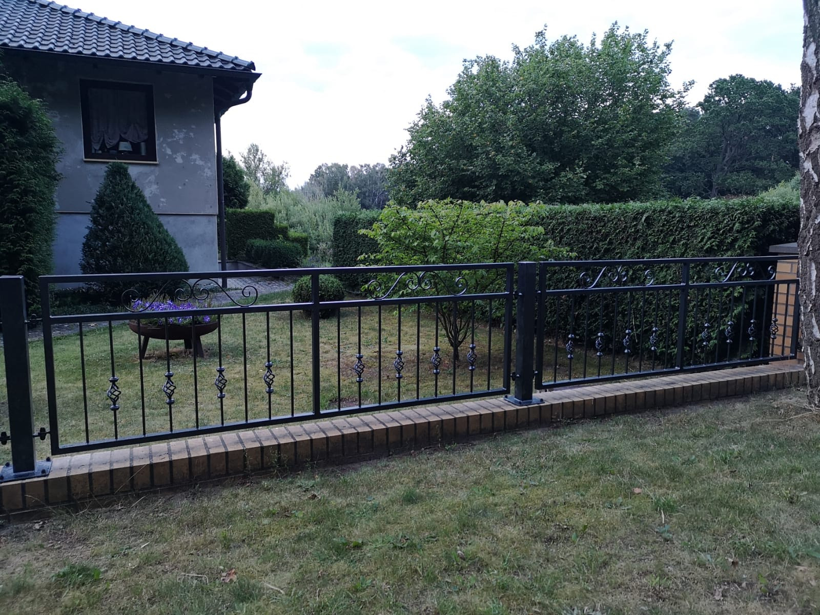 Czarny, metalowy płot z ozdobnymi elementami, posadowiony na niskim murku z cegły klinkierowej, na tle zadbanego ogrodu z domem w tle.