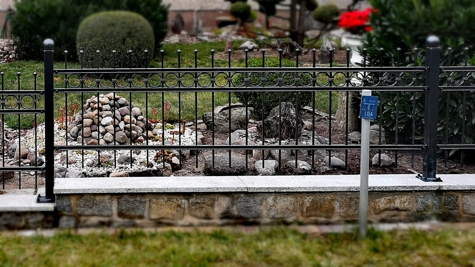 Czarny, metalowy płot z ozdobnymi elementami na tle ogrodu skalnego z kamieniami i zielenią, widoczny słupek z niebieską tabliczką adresową.