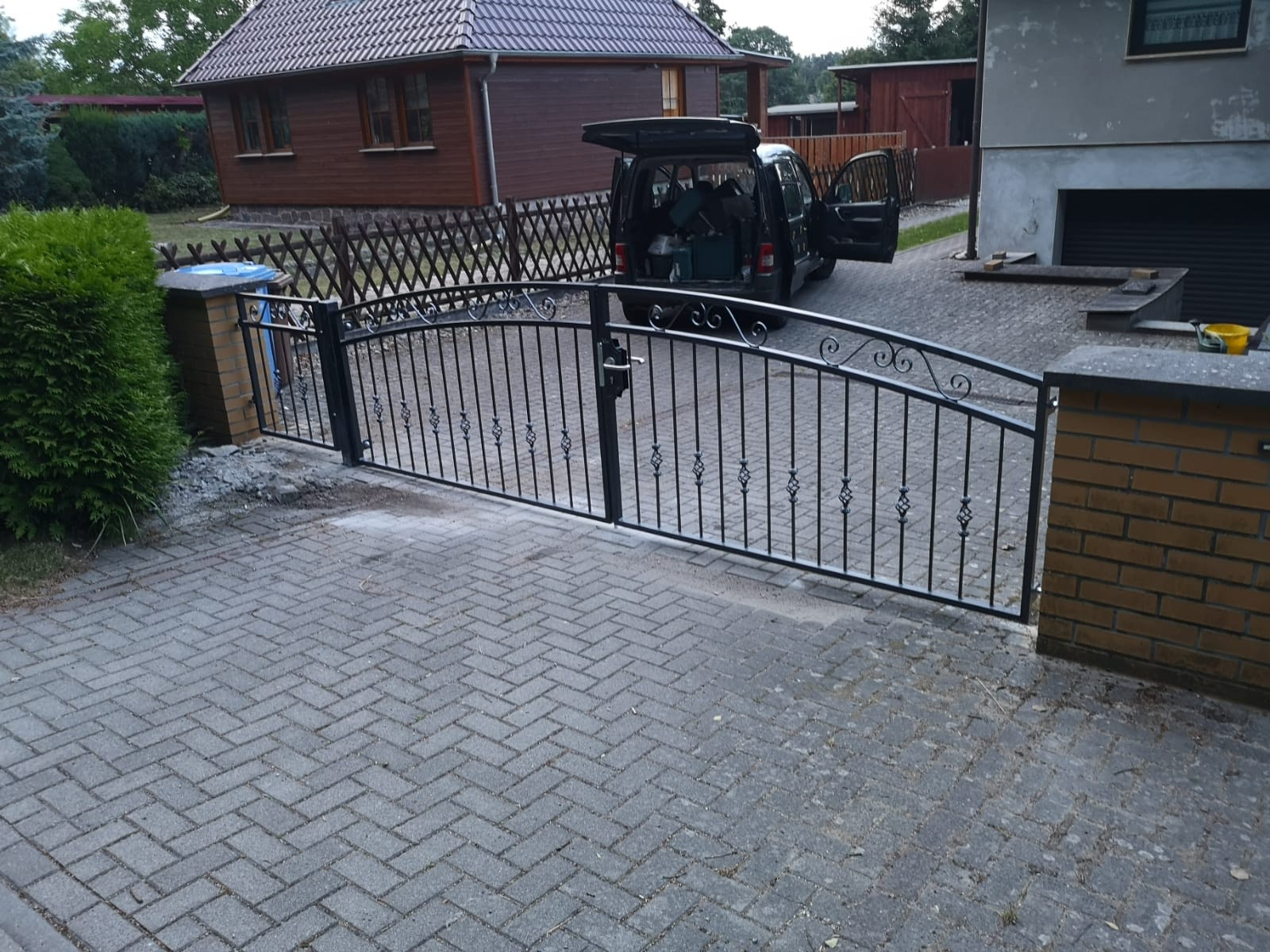Czarna, metalowa brama wjazdowa z ozdobnymi elementami, zamontowana między murowanymi słupkami na posesji z brukowanym podjazdem, samochód z otwartym bagażnikiem w tle.