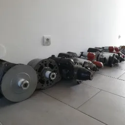 Rząd używanych, zregenerowanych rozruszników samochodowych ułożonych na podłodze warsztatu, widoczny biały kontakt elektryczny na ścianie