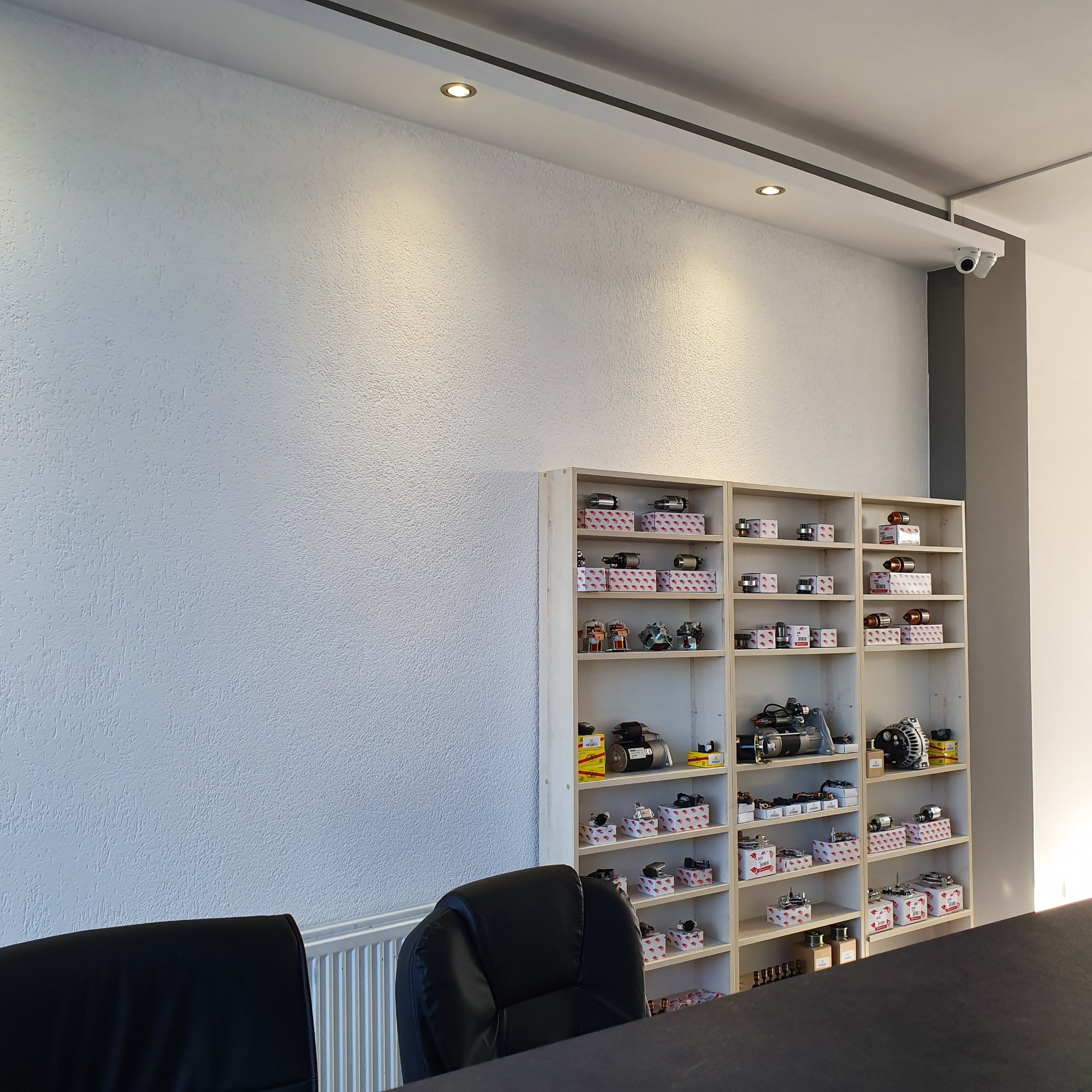 Wnętrze warsztatu samochodowego z regałem wypełnionym częściami zamiennymi, takimi jak rozruszniki, alternatory i inne podzespoły, ułożonymi w pudełkach i luzem na półkach, z widocznymi dwoma...