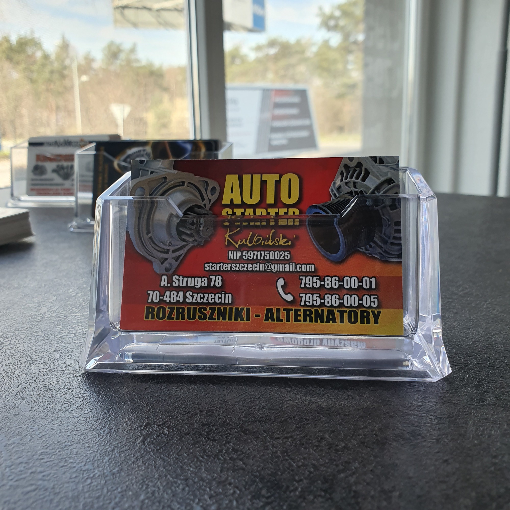 Wizytówka firmy Auto Starter Kulbilski z rozrusznikami i alternatorami w przezroczystym stojaku na biurku.