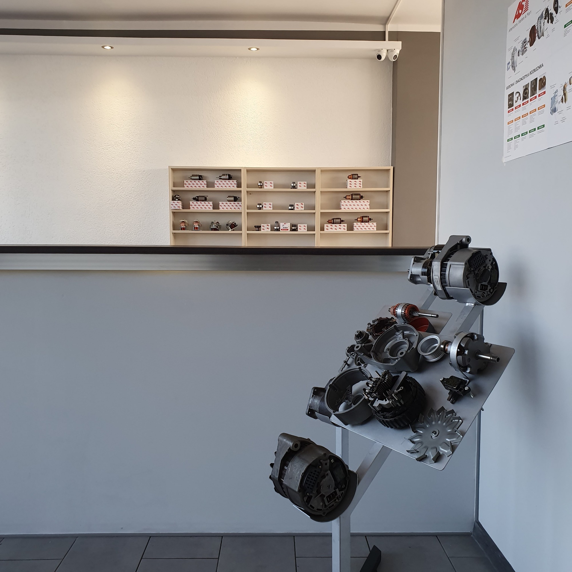 Ekspozycja części samochodowych, w tym alternatorów i rozruszników, na metalowym stojaku w warsztacie, z półkami pełnymi części zamiennych w tle.