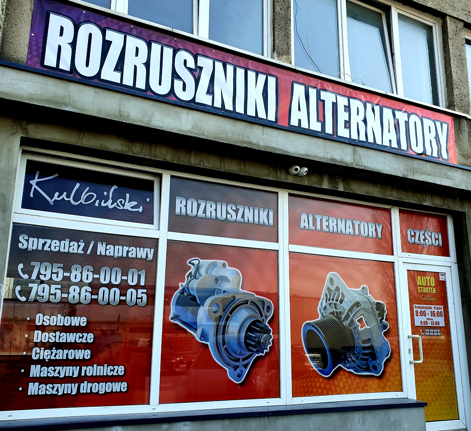 Fasada warsztatu samochodowego specjalizującego się w rozrusznikach i alternatorach, z widocznym banerem i naklejkami na oknach prezentującymi zdjęcia części i dane kontaktowe.