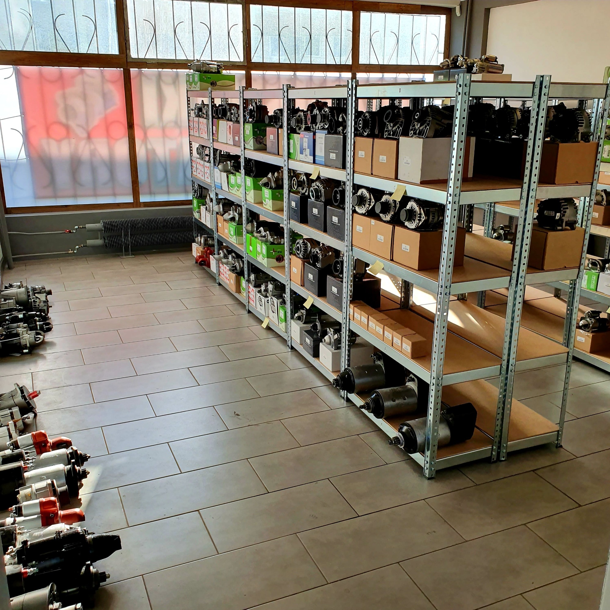 Magazyn części samochodowych: rozruszniki i alternatory ułożone na metalowych regałach, niektóre w pudełkach, inne luzem, z rzędem rozruszników na podłodze w pierwszym planie.
