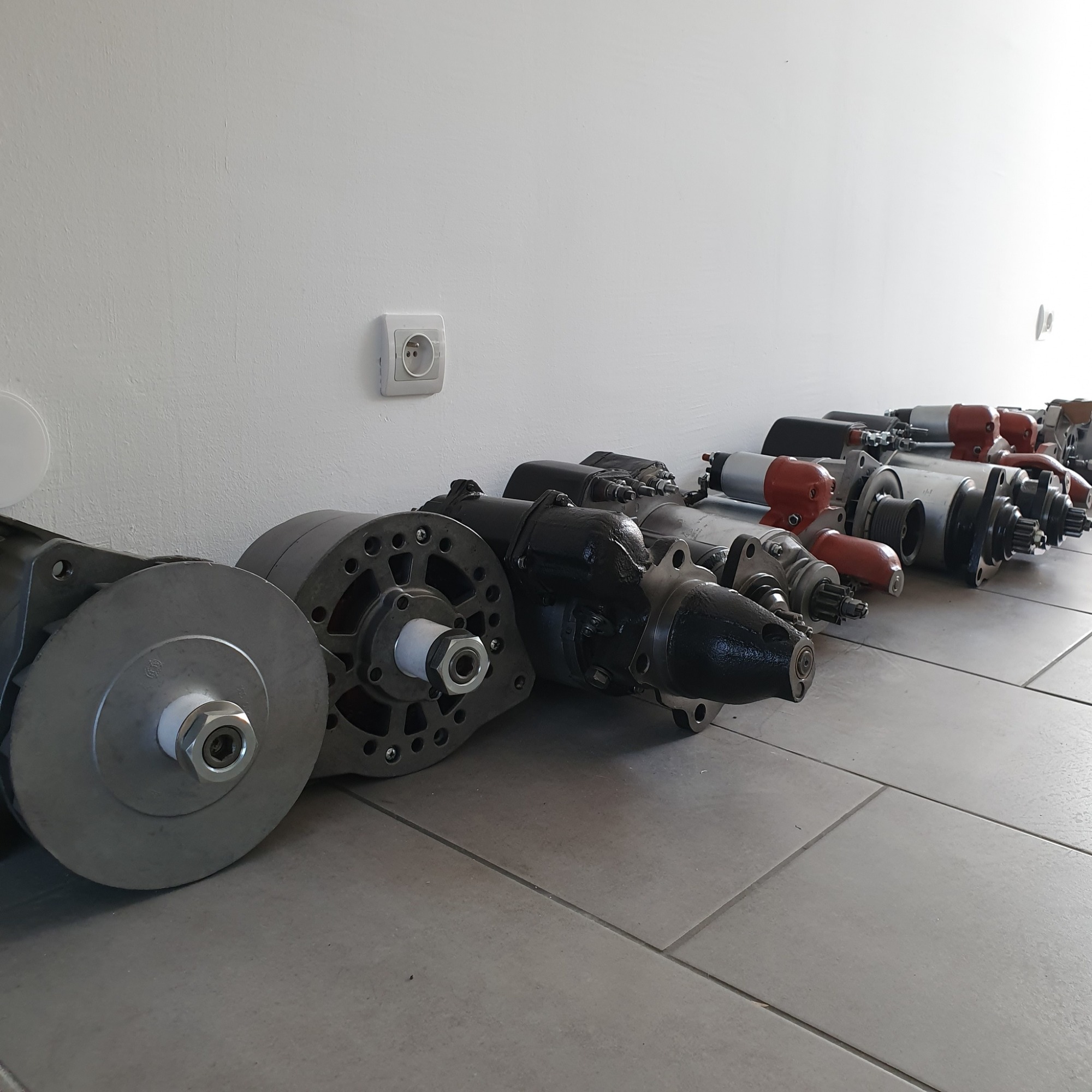 Rząd używanych, zregenerowanych rozruszników samochodowych ułożonych na podłodze warsztatu, widoczny biały kontakt elektryczny na ścianie