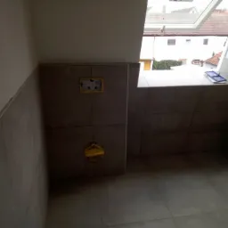 Szara łazienka w trakcie wykańczania z widocznym oknem dachowym, płytki na ścianach i podłodze, żółte elementy instalacji wodnej oraz niebieskie narzędzia na parapecie.