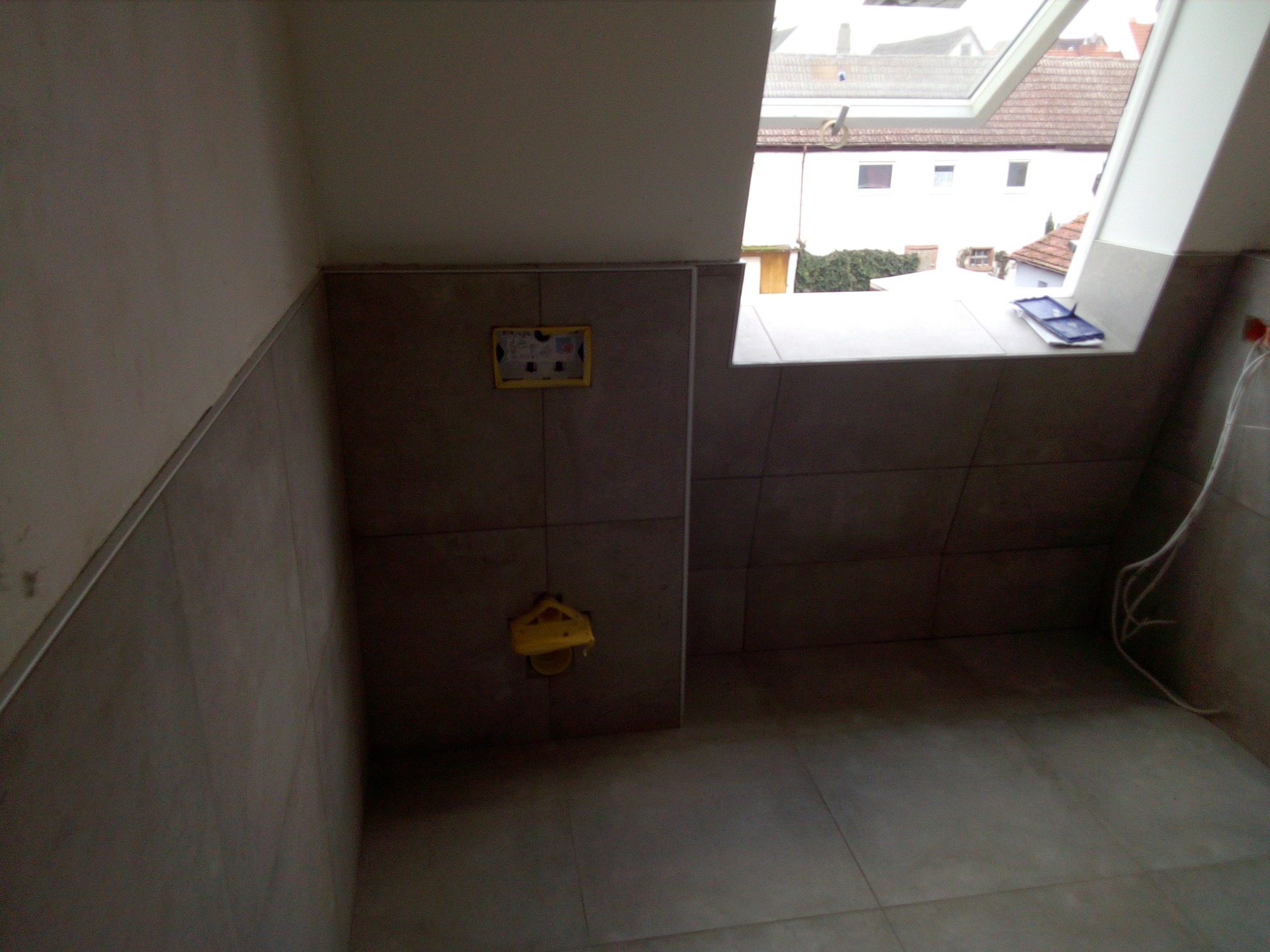 Szara łazienka w trakcie wykańczania z widocznym oknem dachowym, płytki na ścianach i podłodze, żółte elementy instalacji wodnej oraz niebieskie narzędzia na parapecie.