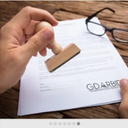 Ręka przykłada drewnianą pieczątkę do umowy leżącej na biurku, obok czarne okulary.