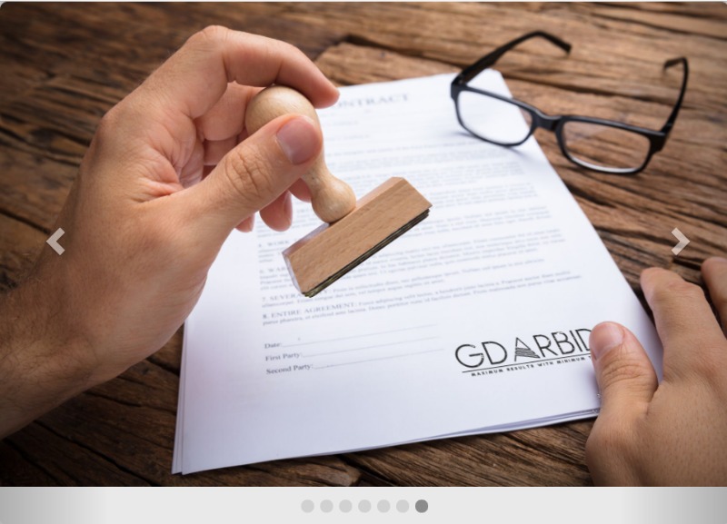 Ręka przykłada drewnianą pieczątkę do umowy leżącej na biurku, obok czarne okulary.