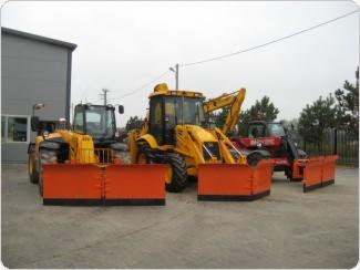 Trzy maszyny budowlane: żółta koparko-ładowarka z pomarańczowym pługiem śnieżnym na pierwszym planie, obok ładowarka teleskopowa i czerwona ładowarka teleskopowa, stojące na placu przed budynkiem.