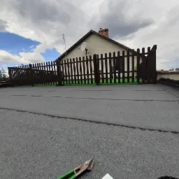 Widok z góry na płaski dach pokryty papą, z niskim, ciemnobrązowym płotkiem otaczającym fragment dachu i domu w tle. Na pierwszym planie leżą nożyce do blachy z zielonymi uchwytami.