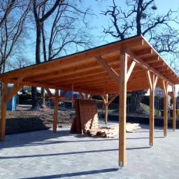 Drewniana wiata z rynnami na posesji, w tle drzewa i błękitne niebo.