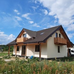 BDM domy Drewniane - Nowoczesny dom z beżową elewacją, drewnianymi balkonami i ciemną dachówką na tle błękitnego nieba z chmurami. Biały samochód dostawczy zaparkowany obok.