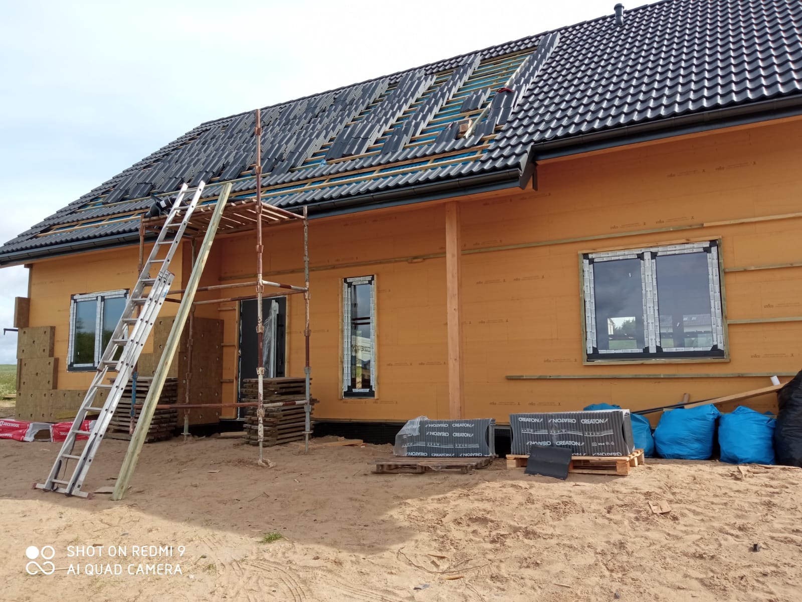 Budowa domu: montaż dachu z dachówki ceramicznej, widoczne rusztowanie, drabina i pakiety dachówek Creaton na paletach przed budynkiem z płyt OSB.
