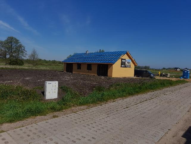 Żółty budynek z niebieskim dachem w trakcie budowy na działce, widoczna skrzynka elektryczna i toaleta przenośna w tle.