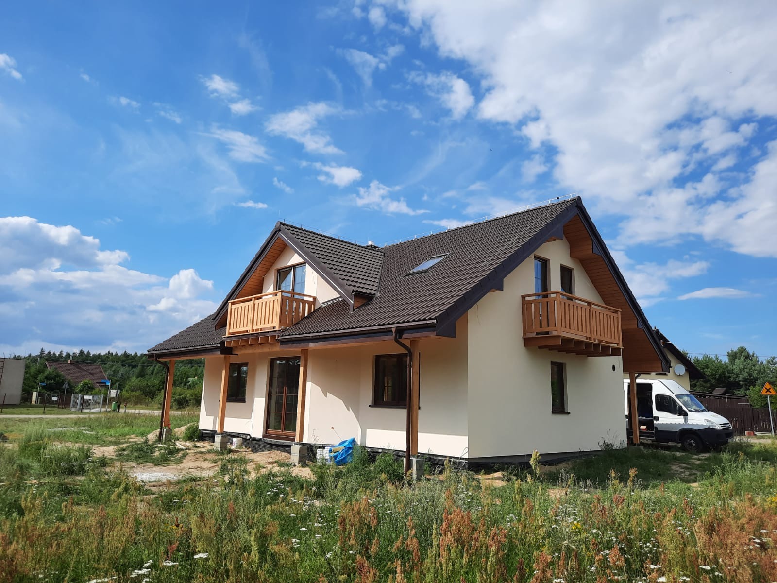 Nowoczesny dom z beżową elewacją, drewnianymi balkonami i ciemną dachówką na tle błękitnego nieba z chmurami. Biały samochód dostawczy zaparkowany obok.