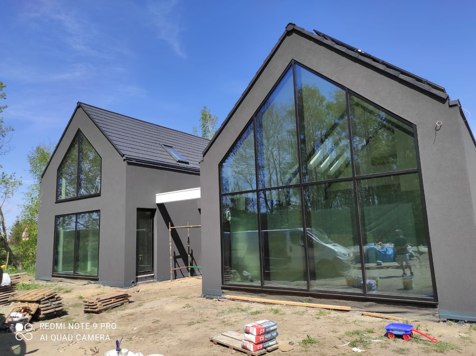 Dwa nowoczesne, szare budynki z dużymi oknami w kształcie trójkąta, widoczne materiały budowlane i narzędzia, błękitne niebo w tle.