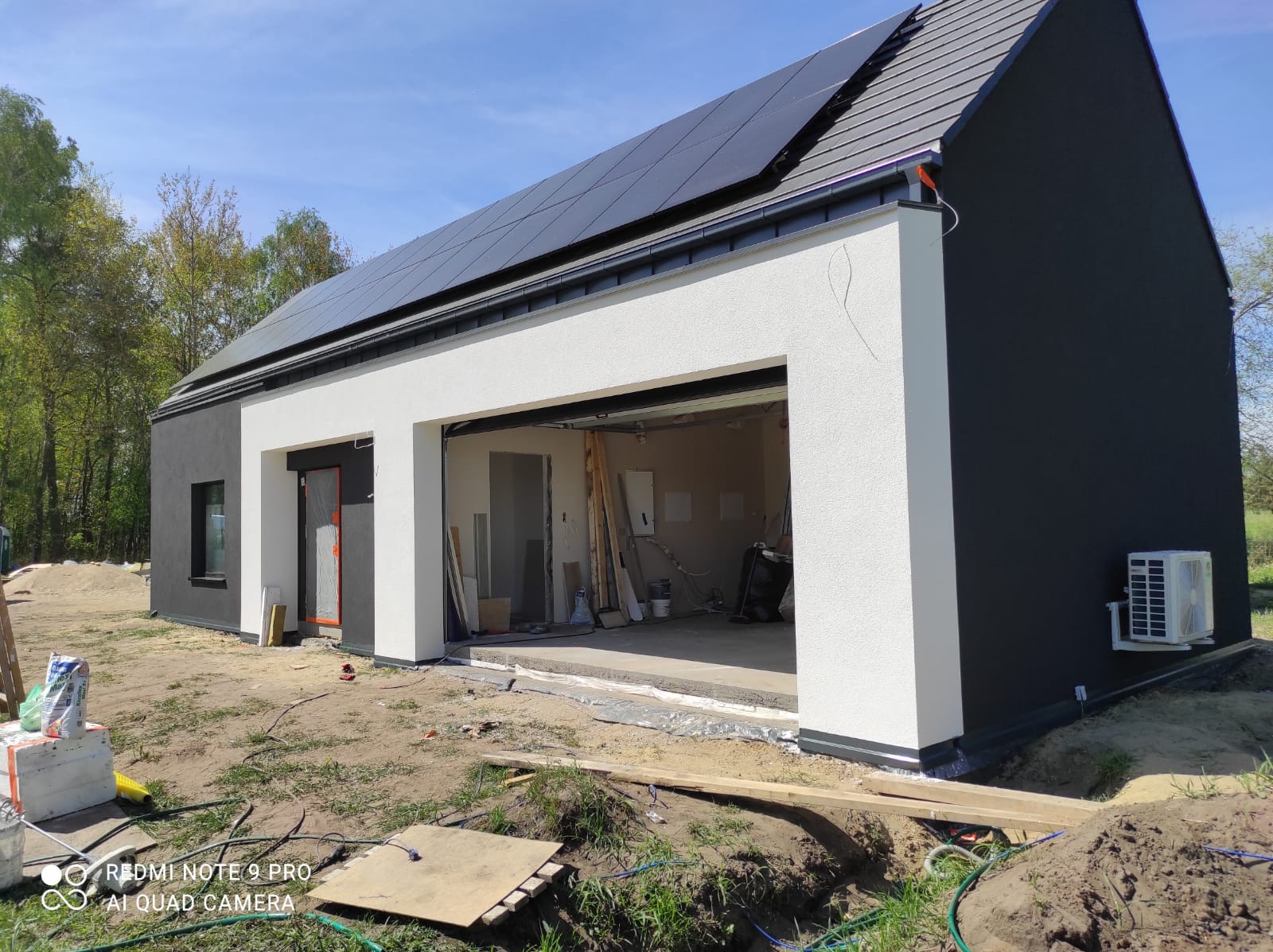 Nowoczesny dom z panelami słonecznymi na dachu w trakcie budowy, widoczny otwarty garaż i klimatyzator na ścianie, teren wokół domu częściowo uporządkowany.