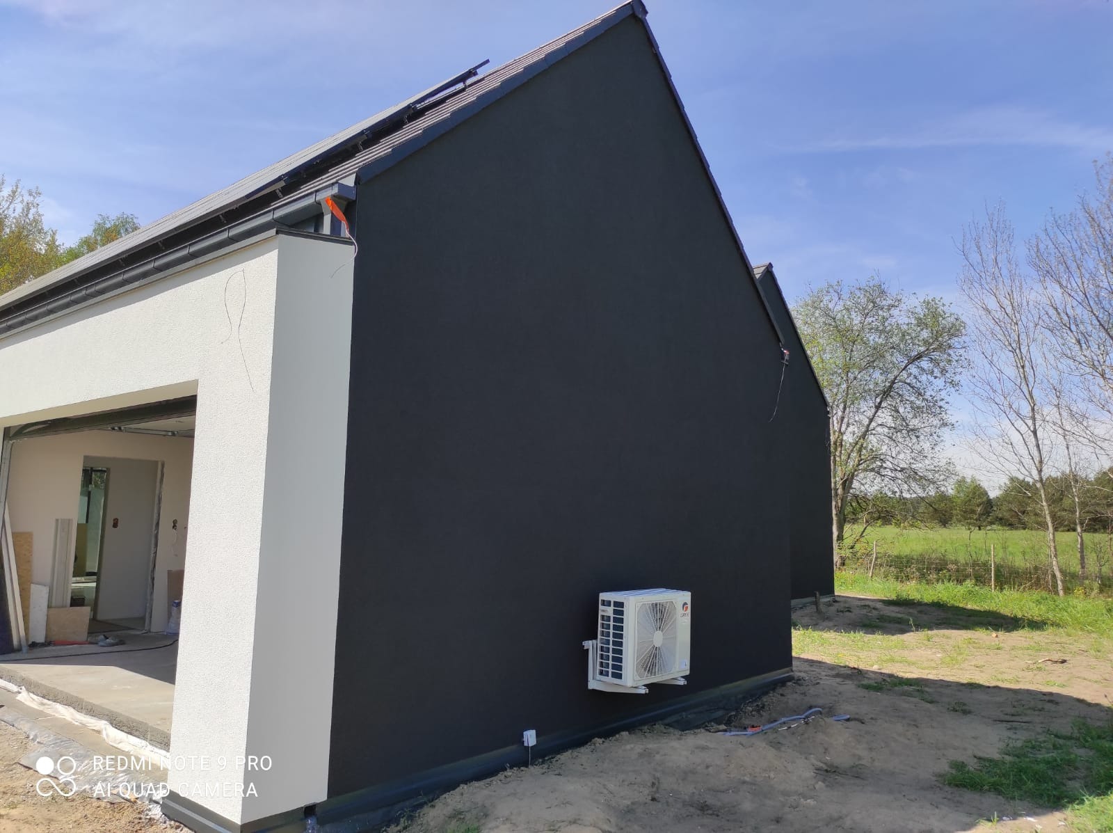 Ciemna elewacja domu z zamontowanym klimatyzatorem, widoczny fragment garażu z surowym wykończeniem i otoczenie z zielenią.